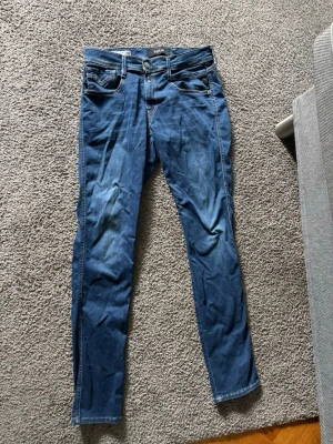 Blå slim fit jeans från Replay - Säljer ett par blå slim fit jeans från Replay, modell Anbass. Jeansen har klassisk femficksdesign, dragkedjegylf och snygga slitningar framtill. Perfekta för dig som gillar en smalare passform och tidlös denimstil.