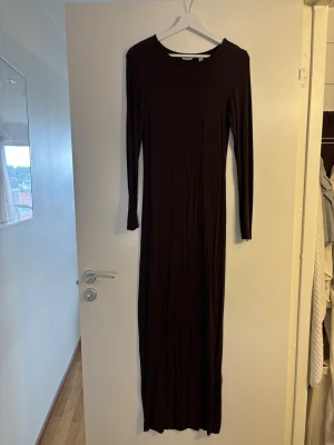 Lång brun klänning med långa ärmar - En stilren lång brun klänning med rund halsringning och långa ärmar. Klänningen har en smal passform och är tillverkad i ett mjukt, stretchigt material som ger en bekväm känsla. Perfekt för dig som gillar minimalistisk och tidlös stil.