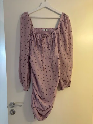 Ljuslila blommig klänning med puffärm - Supersöt ljuslila klänning med små blommor i lila och grönt. Klänningen har långa puffärmar, fyrkantig ringning och smock upptill. Nederdelen har snygg rynkning med dragsko i sidan för en trendig look. Perfekt för dig som gillar romantisk och feminin stil.