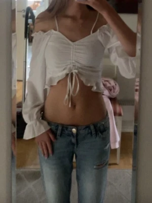 Vit croppad offshoulder blus - Säljer en vit croppad blus med offshoulder och långa ärmar. Toppen har snörning framtill, volangkant nedtill och tunna axelband. Perfekt för dig som vill ha en söt och trendig topp till sommaren.
