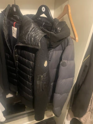 Svart dunväst från Moncler och moncler Madiera - Snygg svart dunväst från Moncler med quiltad design och stickade sidopaneler. Västen har dragkedja framtill, hög krage och Moncler-logga på bröstet. Perfekt för lager-på-lager och håller dig varm med stil. Du får båda jackorna tillsammans 