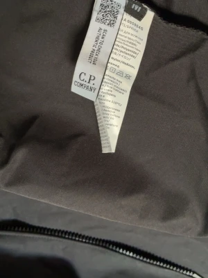 Svart overshirt från C.P. Company - Snygg svart overshirt från C.P. Company med dragkedja framtill, bröstficka och ikonisk linsdetalj på ärmen. Tillverkad i en mix av polyamid och nylon som ger en lätt och modern känsla. Perfekt för lager på lager och streetwear-stil.