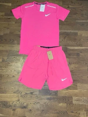 Rosa träningsset från Nike - Säljer ett fräscht rosa träningsset från Nike med t-shirt och shorts. Båda plaggen har den klassiska Nike-loggan i vitt och är tillverkade i ett lätt och snabbtorkande material. Perfekt för gymmet eller löprundan.