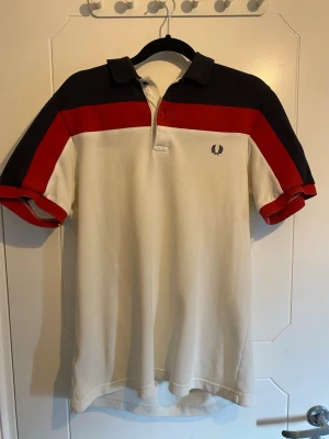 Fred Perry piké med färgblock, M - Säljer en snygg Fred Perry pikétröja i slim fit med färgblock i marinblått, rött och vitt. Tröjan har korta ärmar, klassisk krage och knappslå framtill. Tillverkad i bomull och har Fred Perry-loggan broderad på bröstet.