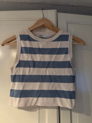 Randig ärmlös topp från Zara - Säljer en snygg ärmlös topp från Zara med breda blå och vita ränder. Toppen har rund halsringning och är croppad i modellen. Perfekt för dig som gillar en fräsch och enkel stil. Materialet känns mjukt och bekvämt.