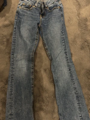 Blå bootcut jeans  - Säljer ett par blå bootcut jeans med snygga kontrastsömmar och detaljerade bakfickor med knapp. Jeansen har klassisk femficksdesign och är tillverkade i denim med lätt tvättad look. Perfekta för dig som gillar en avslappnad men trendig stil.