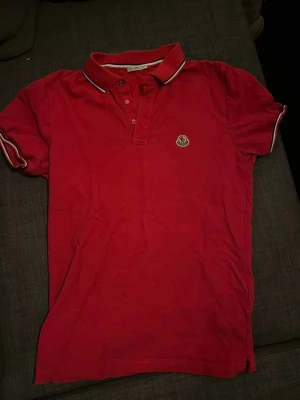 Röd Moncler piké t-shirt - Säljer en röd piké t-shirt från Moncler med klassisk krage och knappar framtill. T-shirten har vita och svarta detaljer på kragen och ärmsluten samt Moncler-logga på bröstet. Perfekt för en stilren och sportig look.