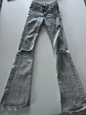 Ljusblå slitna bootcut jeans XS - Säljer ett par ljusblå bootcut jeans med slitningar över knäna och råa, fransiga benslut. Jeansen har klassisk femficksmodell och är tillverkade i denim. Perfekta för en avslappnad och trendig look.