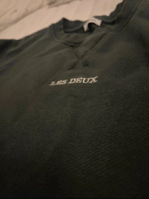Mörkgrön sweatshirt från Les Deux - Mörkgrön sweatshirt från Les Deux med vit broderad logga på bröstet. Tröjan har rund halsringning, ribbade muddar vid ärmslut och nederkant samt en klassisk relaxed passform. Perfekt för en chill och stilren look.