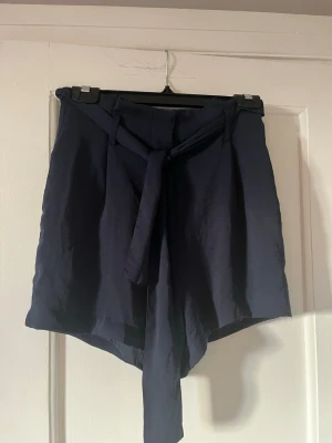 Mörkblå shorts med knytband - Snygga mörkblå shorts med hög midja och ett brett knytband i midjan. De har en lös passform och är tillverkade i ett mjukt tyg som känns skönt mot huden. Perfekta för varma dagar och ger en stilren look.