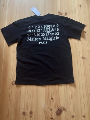Svart Maison Margiela t-shirt - Säljer en svart t-shirt från Maison Margiela med klassisk logga och siffror tryckta på ryggen. Framtill finns de ikoniska vita sömmarna. T-shirten har rund hals och korta ärmar, perfekt för en clean och stilren look.