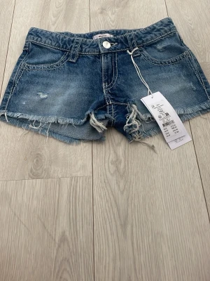Blå jeansshorts med paljettdetaljer - Säljer ett par blå jeansshorts med råa kanter och slitningar framtill. Shortsen har två fickor fram och två bakfickor med snygga paljettdetaljer i silver. Klassisk femficksmodell med knapp och dragkedja. Perfekta för sommaren!