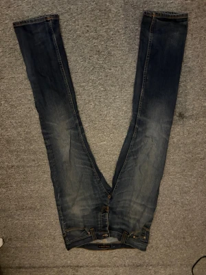 Blå slitna raka jeans - Säljer ett par blå jeans med slitningar och tvättade detaljer. Jeansen har rak passform, klassisk femficksdesign och knappgylf. Materialet är denim i bomull och färgen är mellanblå med ljusare partier på låren och knäna. L 32 w 32
