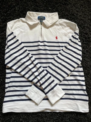 Randig tröja från Polo Ralph Lauren - Vit långärmad tröja med mörkblå horisontella ränder från Polo Ralph Lauren. Tröjan har halv dragkedja, klassisk krage och röd broderad logga på bröstet. Perfekt för en avslappnad men stilren look.