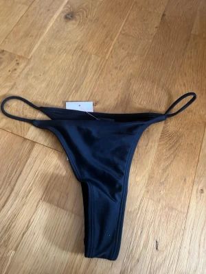 Svart bikiniunderdel  från Nelly - Snygga svarta bikinitroser från Nelly med tunna sidoband och slät, glansig finish. Trosorna har en klassisk stringmodell som ger en minimalistisk look och är tillverkade i ett stretchigt material för skön passform.