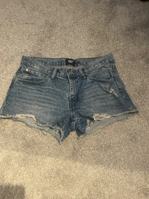 Blå jeansshorts med slitna detaljer - Snygga blå jeansshorts med råa, fransiga kanter och slitna detaljer framtill. Klassisk femficksmodell med knapp och dragkedja. Perfekta för en avslappnad och trendig look i sommar. 