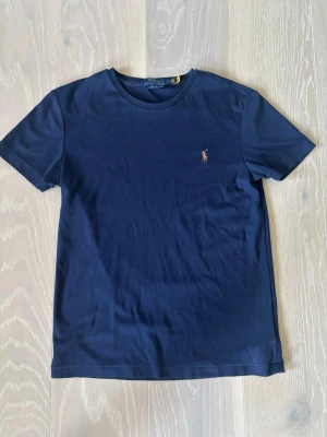 Marinblå t-shirt från Polo Ralph Lauren - Klassisk marinblå t-shirt från Polo Ralph Lauren med broderad logga på bröstet. T-shirten har rund halsringning och korta ärmar. Tillverkad i mjuk bomull för en skön känsla och enkel stil. Vid intresse är det bara att höra av sig