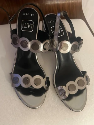 Nya silver Sandaletter i 100 % läder från ILVI Milano - Nytt fint par sandaletter från ILVI Milano i silverfärg i 100 % läder. Sandaletterna är dekorerade med runda metallapplikationer längs remmarna över foten och ankeln. Klacken ( 7,5 cm) är blockformad med en räfflad design. Mycket snyggt par som kommer som kommer att ge en chic känsla till alla outfits vare det vardag eller fest. Inköpt i Italien .  Paret aldrig använts , endast provats.  OBS små i storlek. Det är strl 38, även om det står strl 39 på sandaletterna.  Jag säljer fler fina plagg, sk
