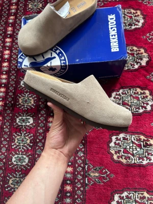 Beige mockatofflor från Birkenstock - Säljer ett par stilrena beige tofflor från Birkenstock i mjuk mocka. De har en klassisk slip-in design med rundad tå och svart sula. Perfekta för dig som vill ha både komfort och enkel stil hemma.