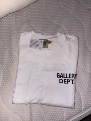 Vit t-shirt från Gallery Dept. - Säljer en clean vit t-shirt från Gallery Dept. med svart logga tryckt på bröstet. Klassisk rund hals och korta ärmar, perfekt till alla outfits. Tillverkad i mjuk bomull för skön känsla hela dagen.