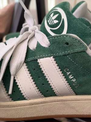 Adidas Campus gröna sneakers - Gröna Adidas Campus sneakers i mocka med vita detaljer och klassiska tre ränder på sidan. Skon har vit sula, breda vita skosnören och Adidas logga på plösen. Snygg retrostil som passar perfekt till streetwear och vardagsoutfits.