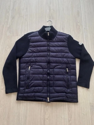 Svart dunväst från Moncler - Snygg svart dunväst från Moncler med quiltad front och stickade ärmar. Västen har två dragkedjeförsedda fickor fram och hög krage. Materialet är en mix av dun och stickad textil, vilket ger en modern och sportig look. Moncler-logga på ärmen för extra stilpoäng.