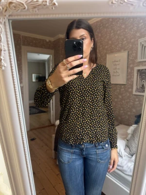 Svart blus med gula prickar - Snygg svart blus med små gula prickar och v-ringning. Blusen har långa ärmar och en avslappnad passform som passar perfekt till jeans eller kjol. Materialet ser ut att vara lätt och skönt, vilket gör den enkel att bära hela dagen.