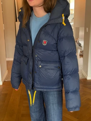 Mörkblå dunjacka från Fjällräven - Snygg mörkblå dunjacka från Fjällräven med huva och gula detaljer på snörningarna. Jackan har två stora fickor framtill med knappar och dragkedja, samt Fjällrävens klassiska logga på bröstet. Perfekt för kalla vinterdagar. Den är i bra skick den har en pytte liten repa på baksidan men inget som är särskilt synligt! Tänk i vinter hur glad du kommer vara även fast det nu är sommar! Den är stor i storleken den är i storlek xxs skulle säga att den är mer i xs eller s då storleken är stor!🩷🩷🩷🩷