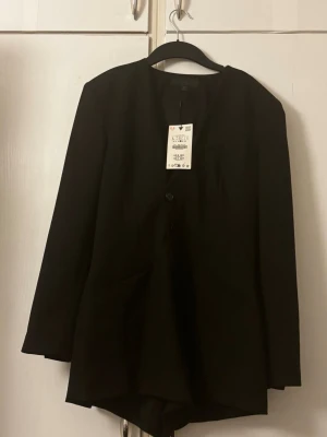 Svart kavaj/jumpsuit - Stilren svart kavaj med rund halsringning och två knappar framtill. Långärmad modell med rak passform och diskreta fickor. Perfekt för en clean och modern look.