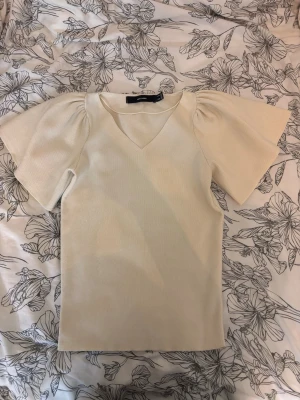 Vit topp med puffärmar och v-ringning - Snygg vit topp med v-ringad hals och puffiga korta ärmar. Materialet är ribbat och är i storlek M men sitter perfekt på mig som bär xs och s. Den är väldigt strechig i materialet för funkar för alla storlekar