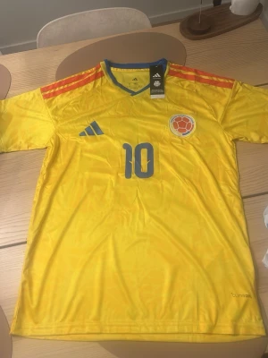 James Rodriguez Colombia Fotbollströj - Säljer en officiell Colombia matchtröja från Adidas i storlek S. Tröjan är gul med blå detaljer, röda ränder på axlarna och nummer 10 på bröstet. Tillverkad i lätt och ventilerande material, perfekt för fotboll eller träning.