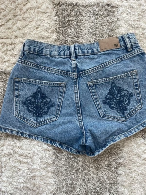 Blå denimshorts med broderade fickor - Säljer ett par klassiska blå denimshorts från 157 med snygga broderade detaljer på bakfickorna. Shortsen har normal passform, fem fickor och knappar  framtill. Perfekta till sommaren och lätta att matcha med allt.