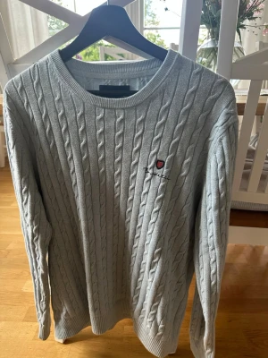 Ljusblå kabelstickad tröja Bondelid - Snygg ljusblå kabelstickad tröja från Bondelid med rund halsringning och broderad logga på bröstet. Den är L men passar M. Tröjan har långa ärmar och ribbade muddar vid ärmslut och nederkant. 