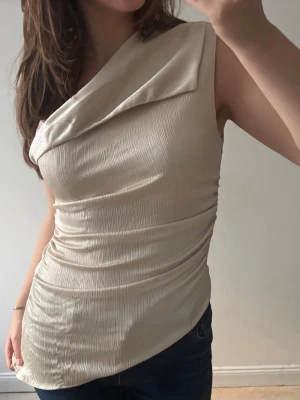 Beige oneshoulder topp i plisserat tyg - Säljer en beige oneshoulder topp med plisserad struktur och asymmetrisk krage. Toppen är figurnära och har ett snyggt fall över axeln. Perfekt för dig som vill ha en unik och trendig look.