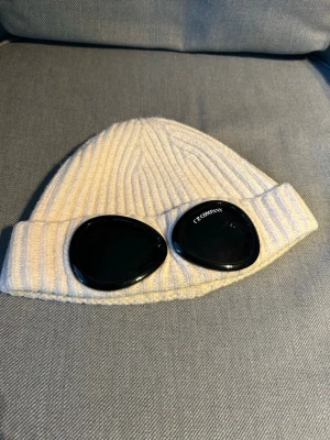 Cp company mössa - Säljer en vit ribbstickad mössa med svarta goggles framtill. Mössan har uppvikt kant och är tillverkad i ett mjukt stickat material. Perfekt accessoar för att sticka ut i mängden och hålla sig varm under kalla dagar. Liten i storleken, kvitto medföljer