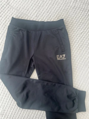 Emporio Armani Svarta EA7 mjukisbyxor - Svarta mjukisbyxor från EA7 Emporio Armani med resår i midjan och muddar vid bensluten. Byxorna har två sidofickor och en vit EA7-logga på ena benet. Perfekta för en avslappnad och sportig stil.