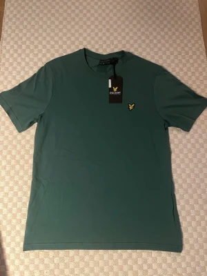 Grön t-shirt från Lyle & Scott - Snygg grön t-shirt från Lyle & Scott med klassisk rund hals och korta ärmar. På bröstet sitter den ikoniska gula loggan. Tillverkad i mjuk bomull för skön känsla och enkel stil. Perfekt till jeans eller shorts.