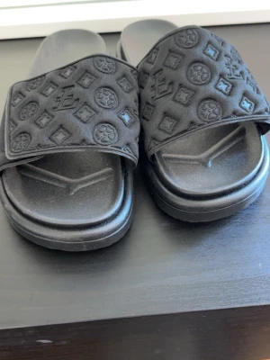Svarta slides med monogramdetaljer - Säljer ett par svarta slides med breda band över foten, dekorerade med upphöjda monogram och mönster. Sandalerna har en chunky sula och är tillverkade i en kombination av textil och syntetmaterial. Perfekta för en avslappnad och trendig look. Aldrig använda. 