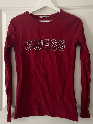 Vinröd långärmad topp från Guess - Snygg vinröd långärmad topp från Guess med rund halsringning och stora bokstäver i kristaller framtill. Använd 2 gånger. 