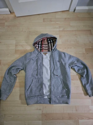 Burrbery hoodie (Helt ny) - Snygg grå hoodie med dragkedja och två fickor framtill. Insidan av huvan och öppningen har ett klassiskt beige, svart och rött rutmönster. Tillverkad i mjukt material och har ribbade muddar vid ärmar och nederkant. Perfekt för en avslappnad stil. Hellt ny är M men känns som S typ och är äkta.. Priset kan diskuterad