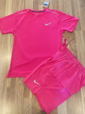 Rosa träningsset från Nike - Säljer ett sportigt träningsset från Nike i stark rosa färg. Setet består av en t-shirt med rund hals och korta ärmar samt matchande shorts med resår i midjan. Båda plaggen har Nike-logga i vitt. Perfekt för gymmet eller löprundan.