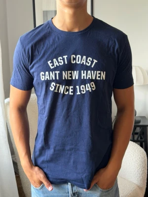 Marinblå Gant t-shirt med tryck - Snygg marinblå t-shirt från Gant med vitt tryck på bröstet där det står 'East Coast Gant New Haven Since 1949'. Klassisk passform och rund halsringning. Perfekt till jeans eller shorts för en avslappnad look.