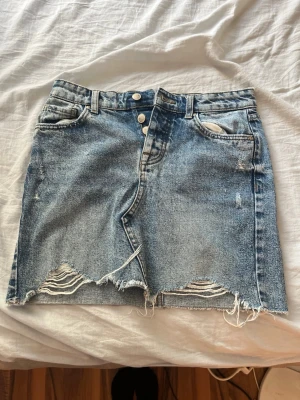 LjusBlå jeanskjol med slitna detaljer från only. - Säljer en blå jeanskjol från ONLY med slitna detaljer och rå kant nertill. Kjolen har knappgylf, fem fickor och en kort passform. Perfekt för dig som gillar en avslappnad och trendig stil.