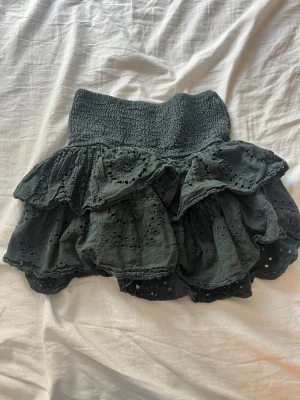 Mörkgrå volangkjol i bomull. Från meet me there. - Supersöt mörkgrå kortkjol med bred smock i midjan och flera lager volanger. Kjolen har broderade detaljer och är gjord i mjuk bomull, perfekt för sommarens alla outfits.