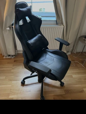 Svart gamingstol med justerbara armstöd - Säljer en svart gamingstol i konstläder med hög rygg, nackkudde och svankstöd. Stolen har hjul, justerbara armstöd och ergonomisk design för extra komfort under långa spelsessioner. Perfekt till skrivbordet eller gaminghörnan.  Den får hämtas om du vill ha den frakt finns inte..
