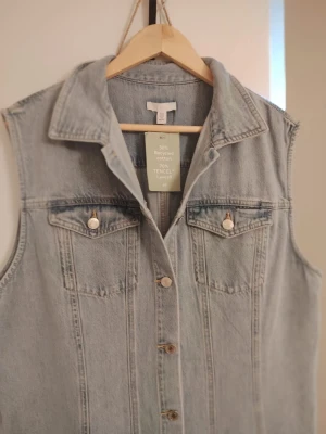 Lång ärmlös jeansklänning - Snygg lång jeansklänning i ljusblå denim med knappar hela vägen framtill och två bröstfickor med lock. Klänningen är ärmlös och har klassisk krage, perfekt för en avslappnad och trendig look.