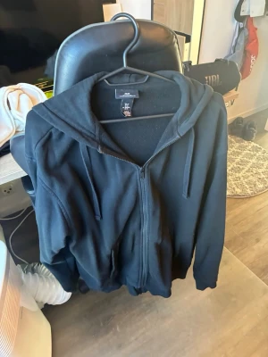 Svart hoodie med dragkedja från H&M - En svart hoodie från H&M i relaxed fit med dragkedja framtill och justerbar huva med snören. Tillverkad i mjukt material, perfekt för chill dagar. Klassisk look med fickor på sidorna och ribbade muddar vid ärmslut och nederkant.