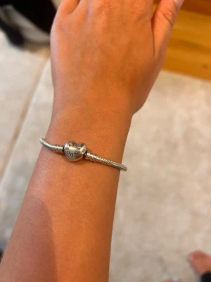 Silverfärgat armband med hjärtlås - Ett ganska nytt Pandora armband som knappast är andvänt, min handled är 16 cm och armbandet är 18 cm det sitter lite stort på mig men det åker inte av älskar värkligen armbandet men har 2.             