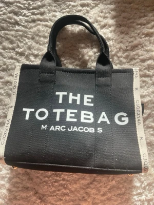 Svart tote bag - Säljer en helt oanvänd svart handväska från en marknad i Spanien med texten 'THE TOTE BAG' i vitt på framsidan. Väskan har vita detaljer på sidorna och har dubbla handtag. Man får med ett längre band. Den är rymlig och har flera fack (se de två sista bilderna). Perfekt för dig som vill ha en trendig och rymlig tote med cool design. INTE ÄKTA!!