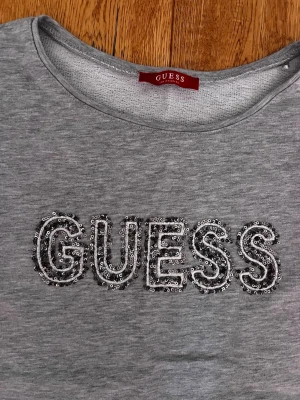 Grå sweatshirt från Guess med paljetter - Snygg grå sweatshirt från Guess med rund halsringning och långa ärmar. Framsidan har ett stort GUESS-tryck dekorerat med glittriga paljetter. Tillverkad i mjukt material som känns skönt mot huden. Perfekt för en avslappnad och trendig look.